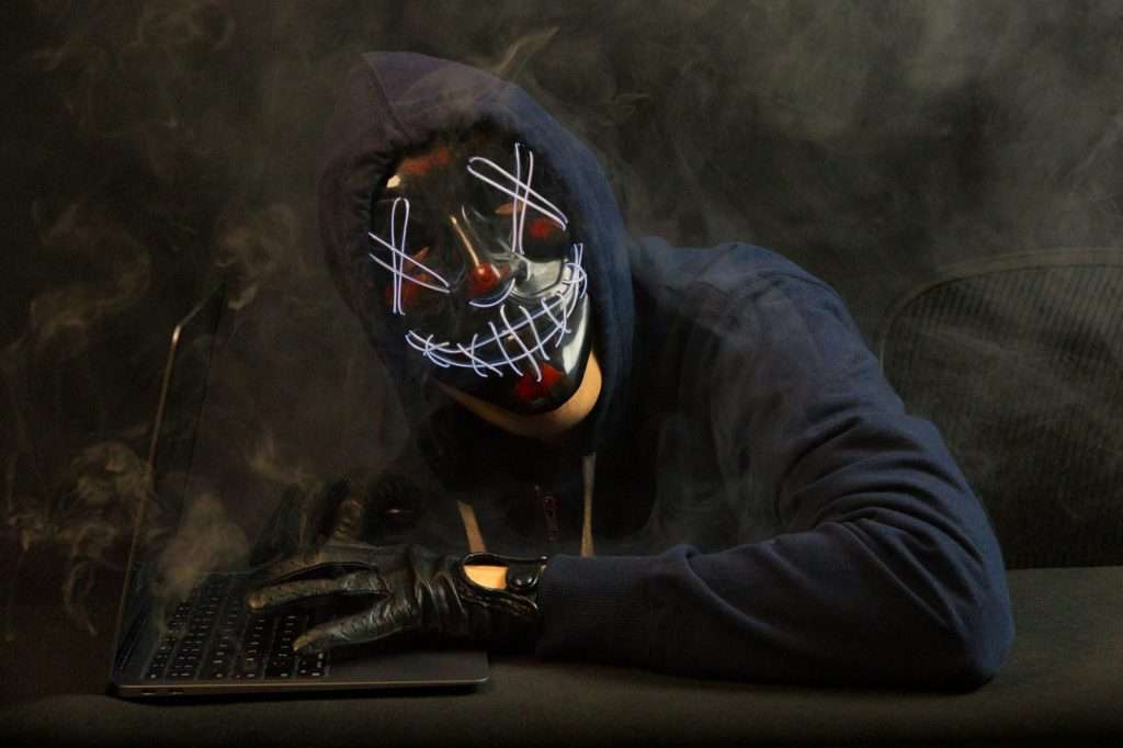 Hooded hacker wearing a glowing red purge mask typing on a laptop in the dark — AI-driven cyberattacks against small businesses are real and happening now. Photo by Kaptured by Kasia / Unsplash.